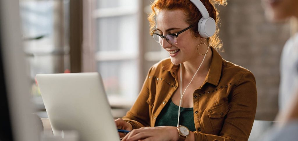 image of a red-headed woman wearing glasses working on a computer while wearing headphones