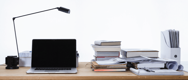 Image of a desk with a lamp, laptop, and various books and papers on it.
