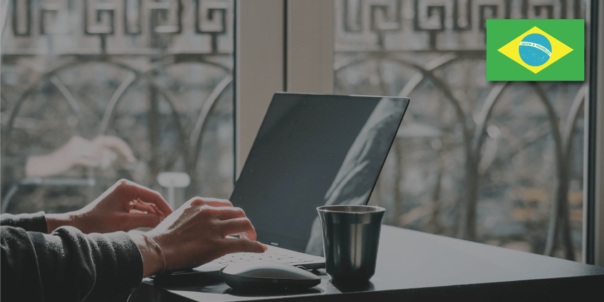 Hands typing on laptop next to mug on desk, Brazil flag icon overlay